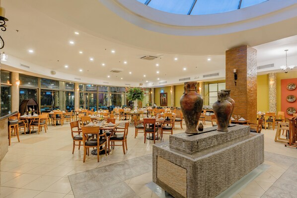 A spacious indoor dining area with wooden tables and chairs, a central decorative stone structure, and large windows allowing natural light. - Holiday Inn Managua - Convention Center by IHG (Managua)