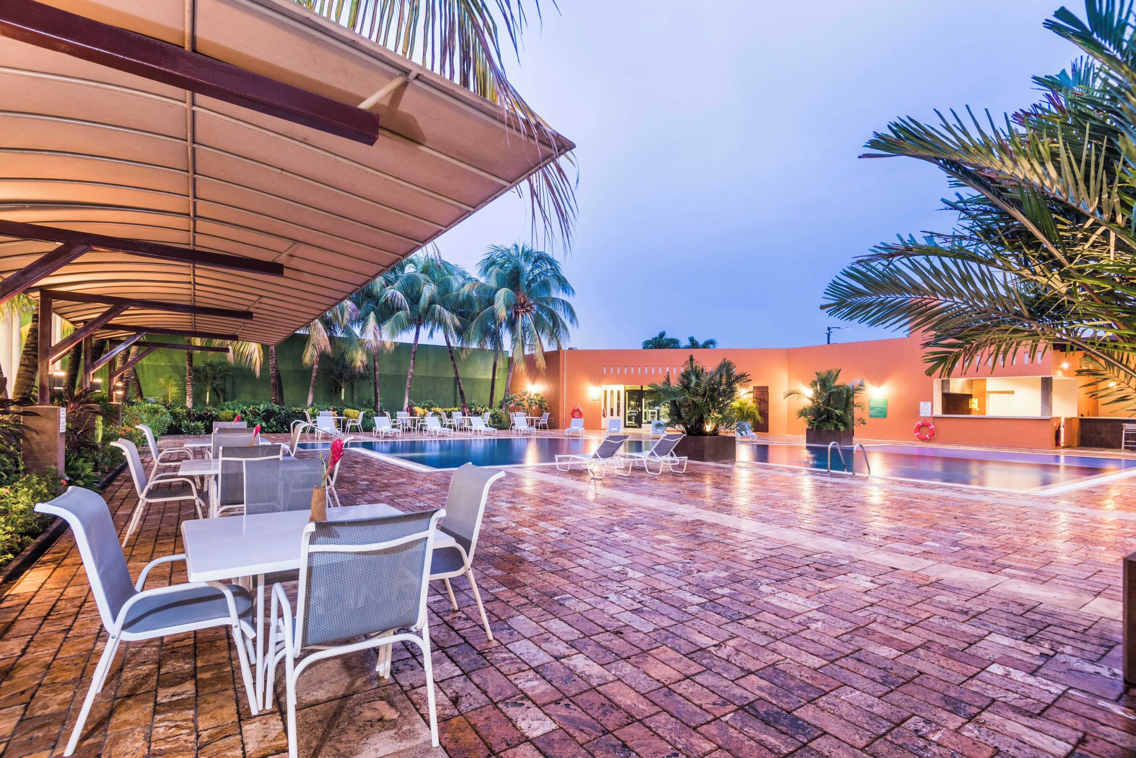 An outdoor pool area with lounge chairs and tables, surrounded by palm trees and a building with an orange facade.