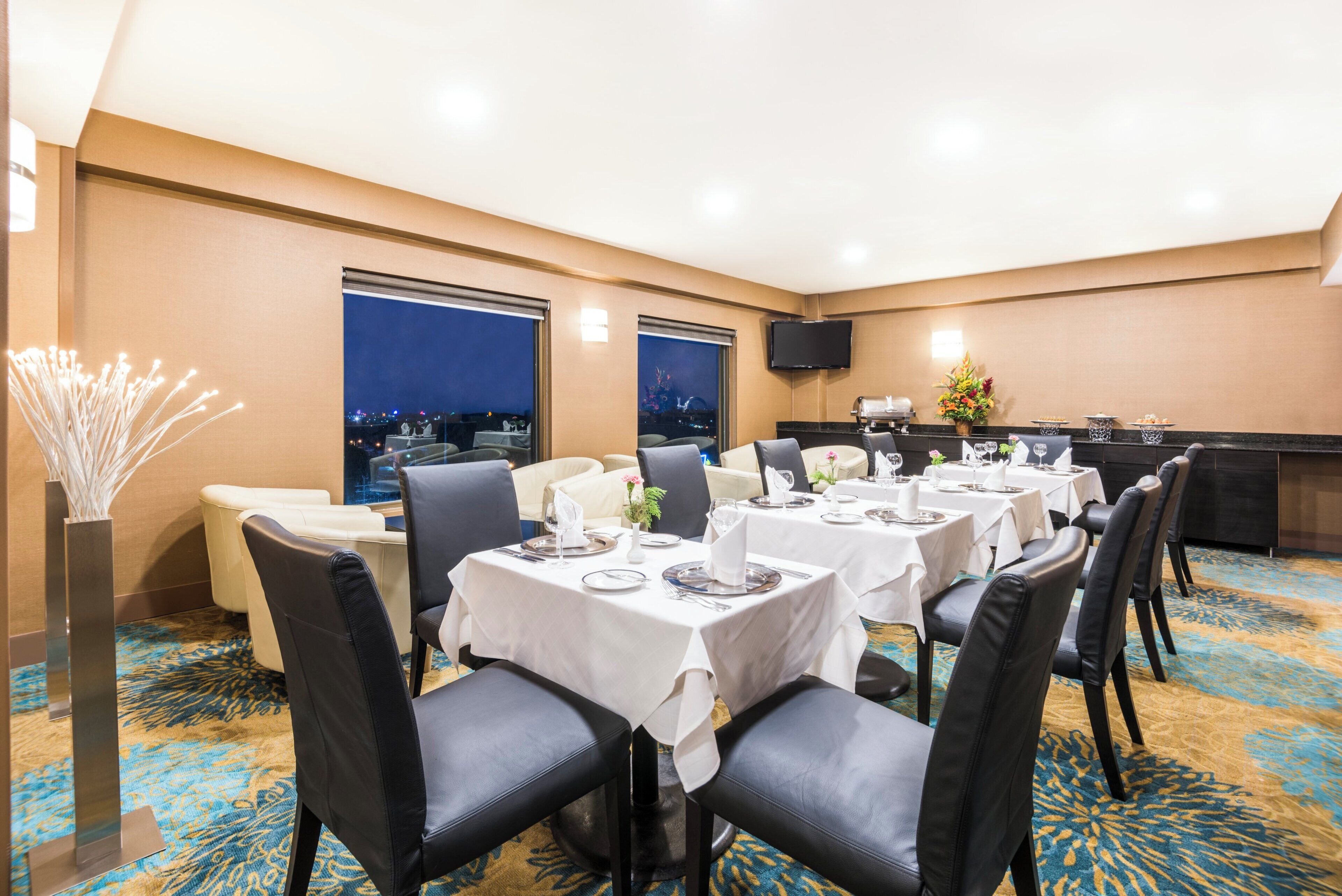 Ein Restaurantbereich eines Hotels mit gedeckten Tischen, einem an der Wand befestigten Flachbildfernseher und Blick auf die Stadt durch die Fenster.