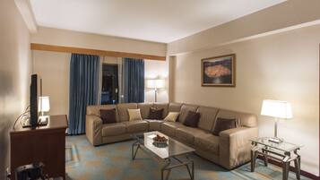 A hotel room with a beige sofa, a glass coffee table, a TV, and a lamp.