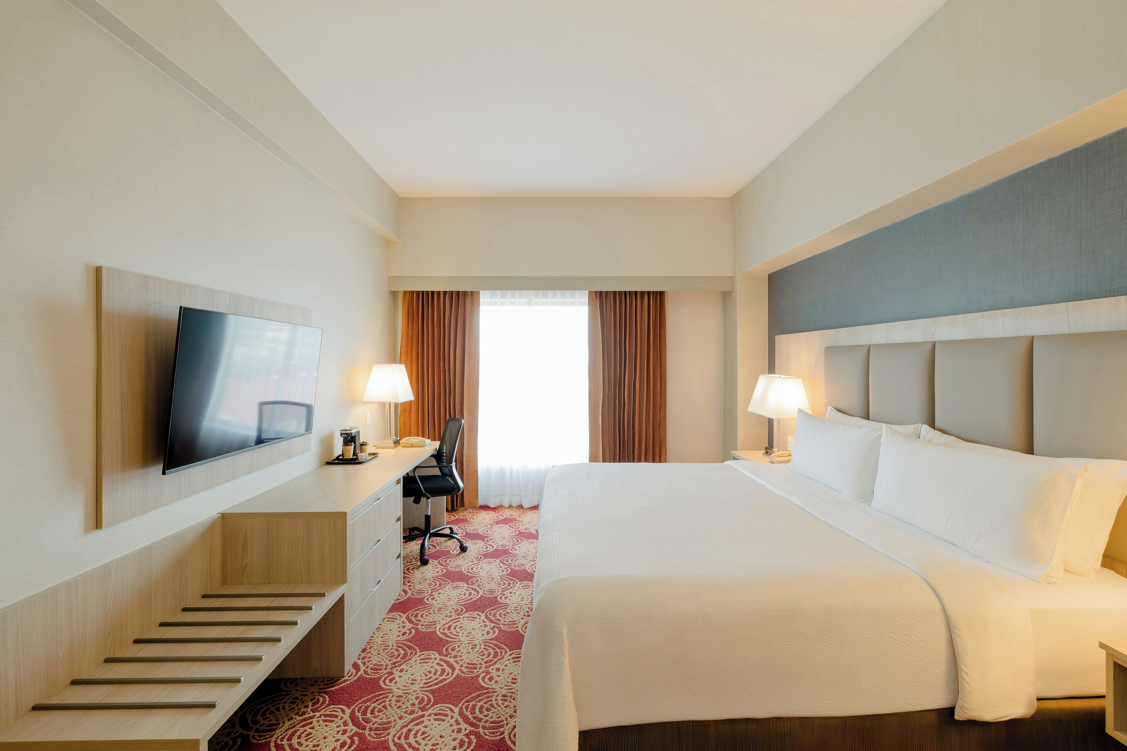 Ein Hotelzimmer mit Bett, Schreibtisch, Stuhl, Fernseher und einem Fenster mit Vorhängen.