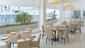 A modern dining area with round tables, beige chairs, and large windows overlooking a pool and outdoor area. - Harlington Ashkelon (Ashkelon)