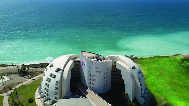 A modern hotel with a unique architectural design featuring two large, curved structures and a central tower. The hotel is located near a beach with a view of the ocean. - Harlington Ashkelon (Ashkelon)