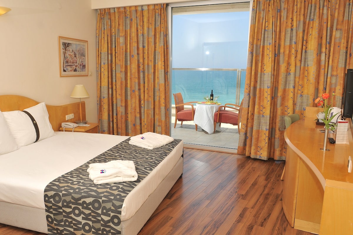 A hotel room with a bed, a desk, a chair, a table with a bottle and glasses, and a view of the sea.