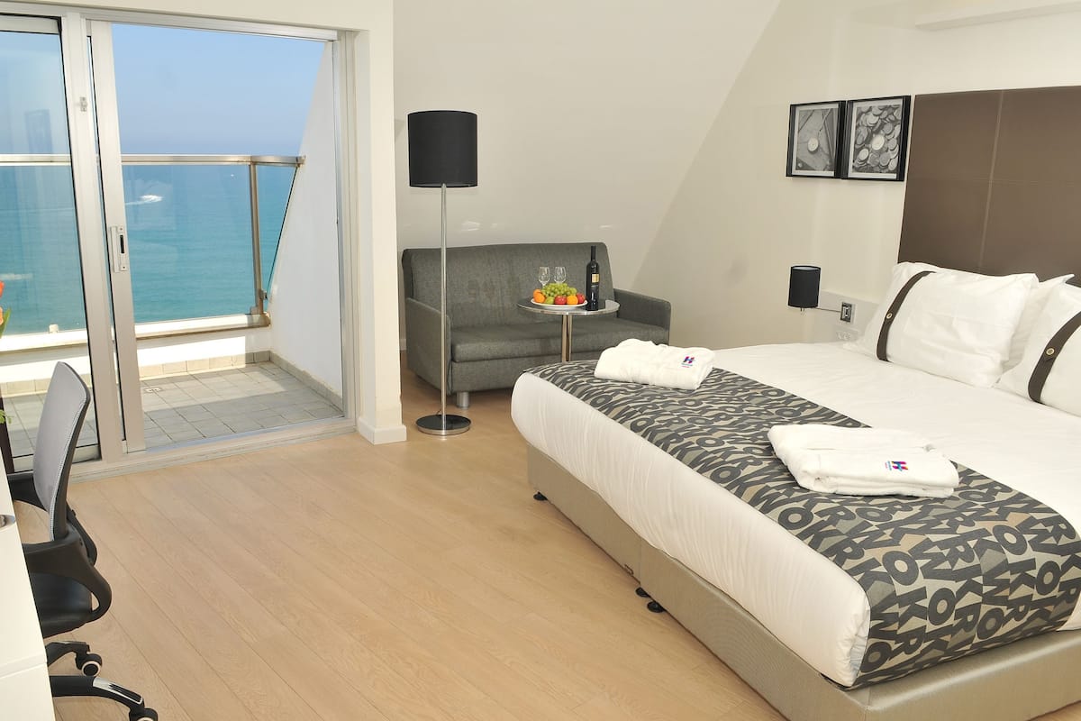 A hotel room with a bed, a sofa, a small table with fruit and wine, a lamp, and a view of the sea.