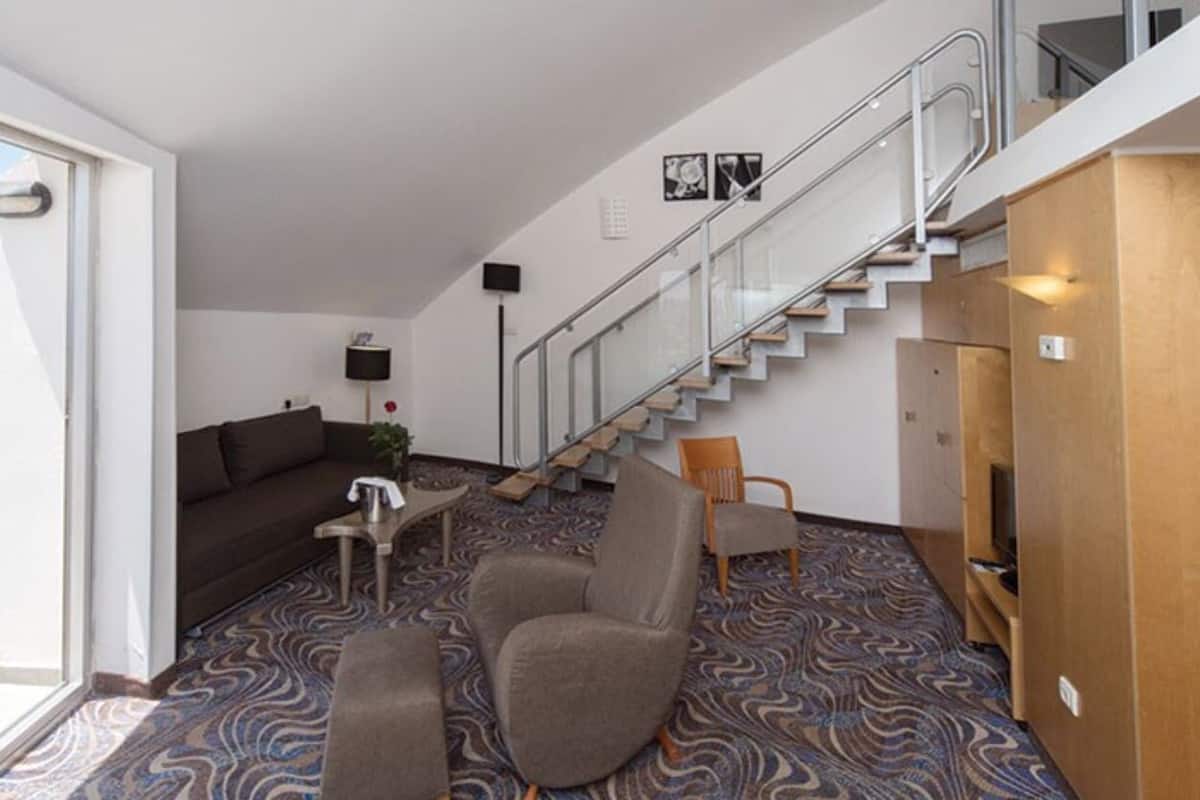 A modern hotel room with a sofa, armchairs, a small table, and a staircase leading to an upper level.
