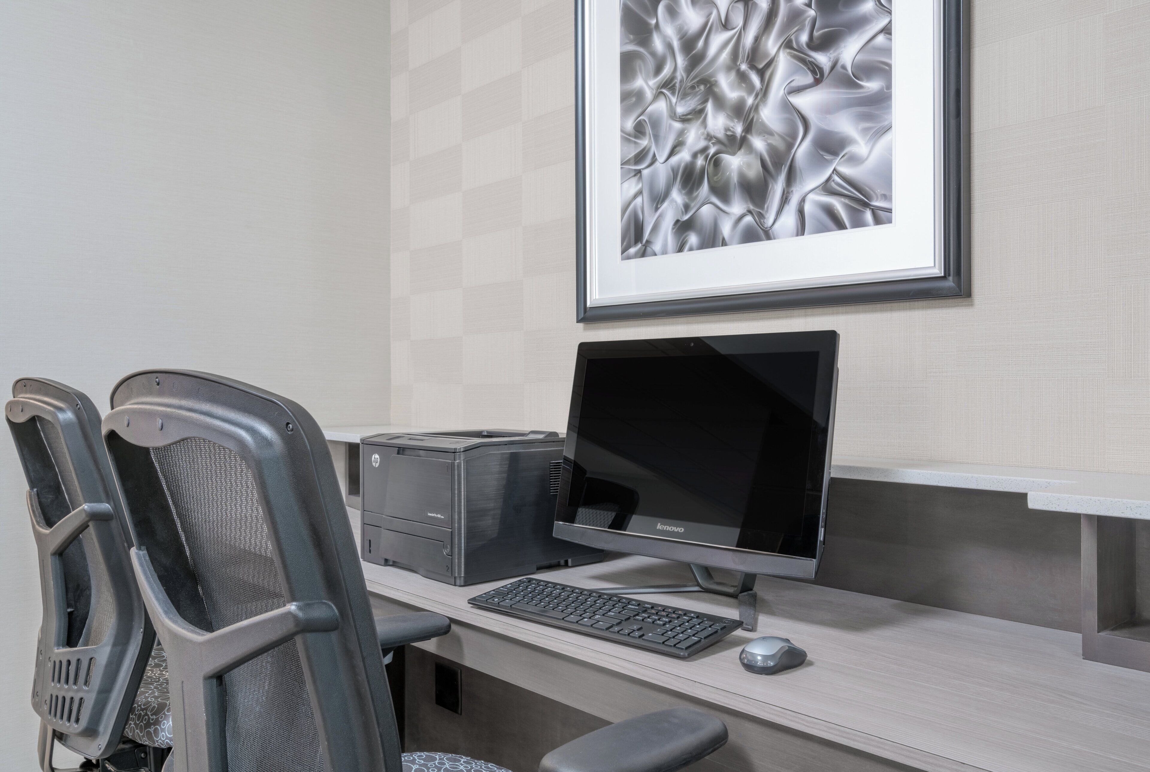 A modern office space with a desktop computer, two office chairs, a framed artwork on the wall, and a printer.