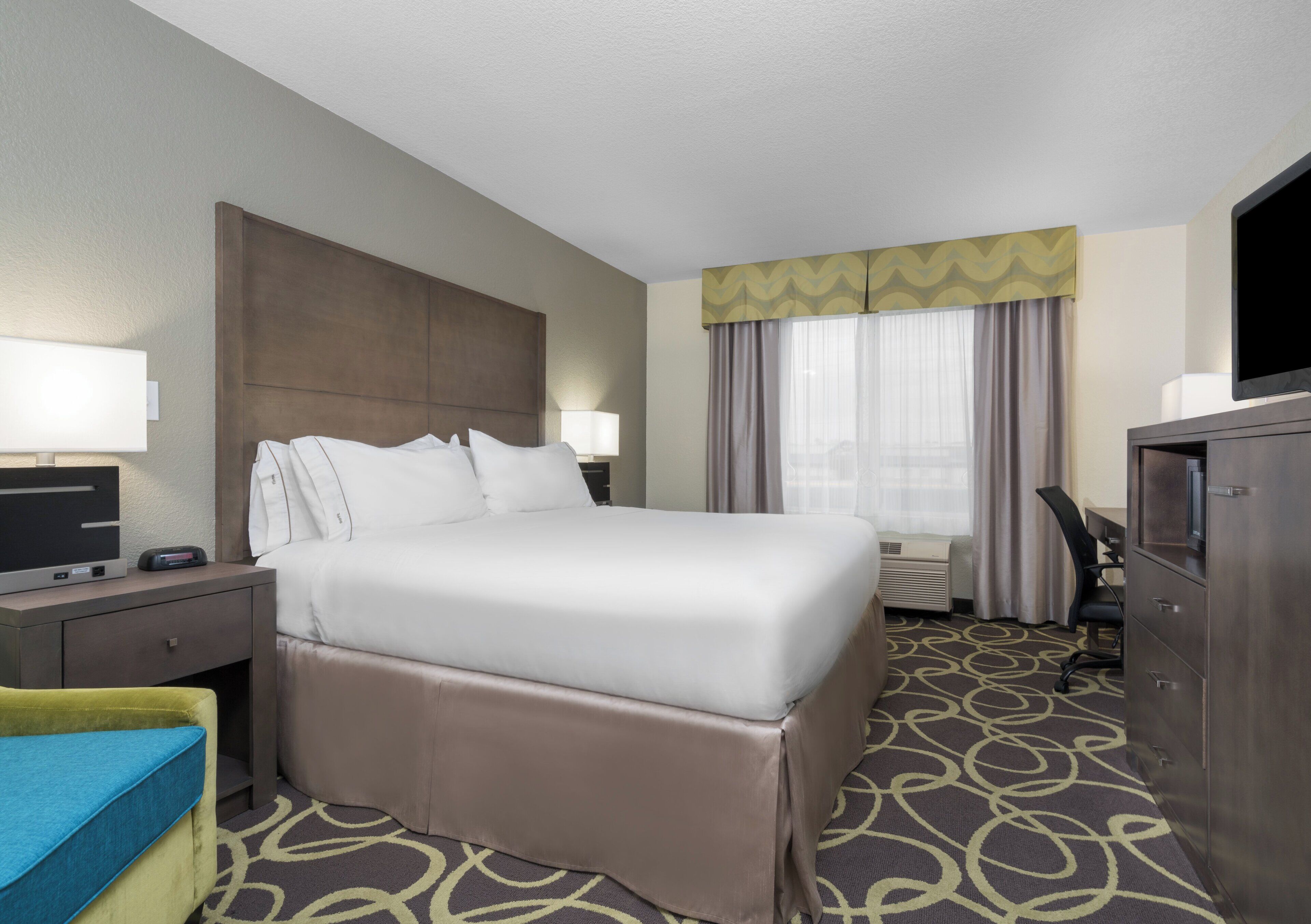 A hotel room with a bed, bedside tables, a desk, a television, and a patterned carpet.