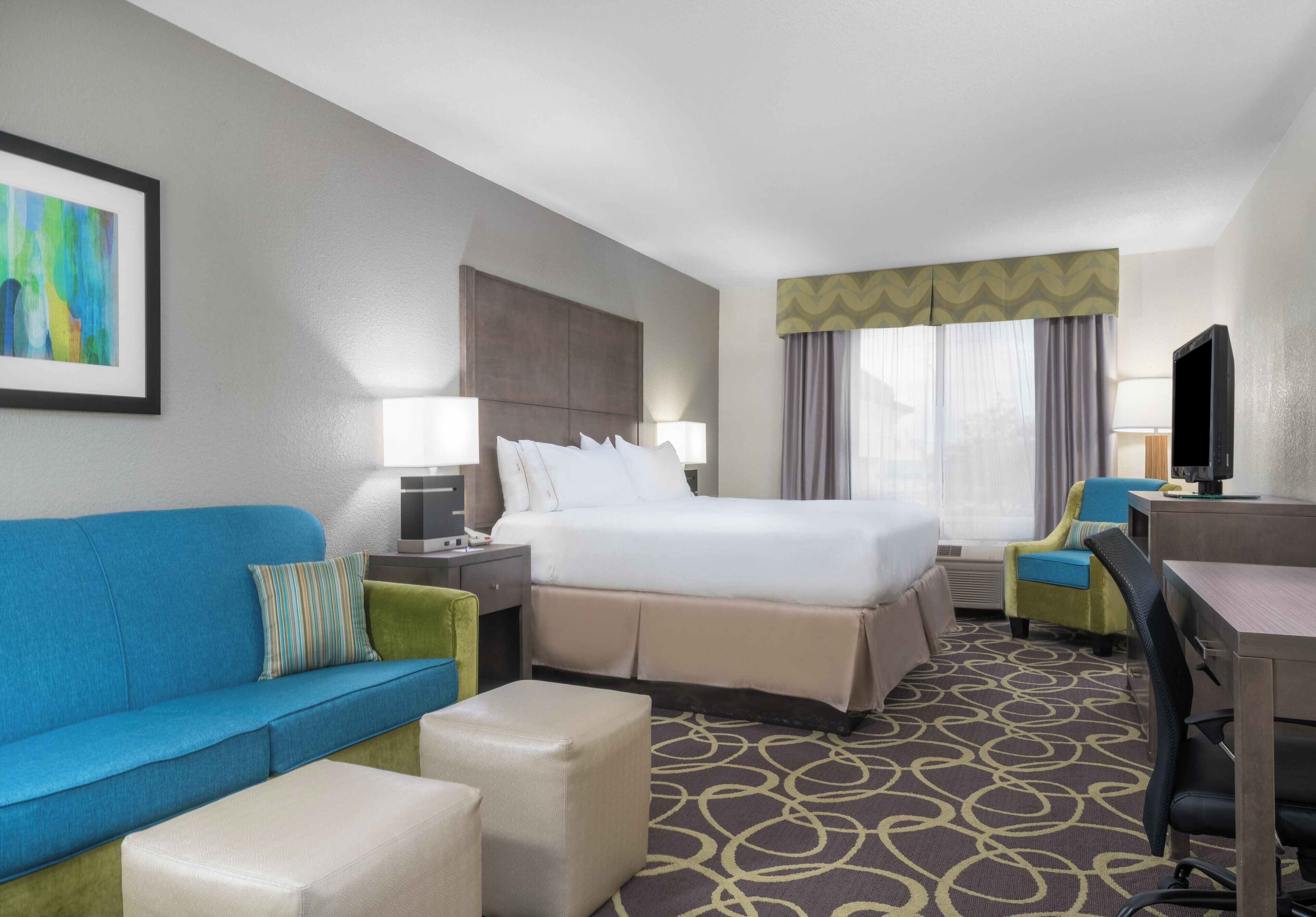 A hotel room with a bed, a blue sofa, a green armchair, a desk with a computer, and a television.
