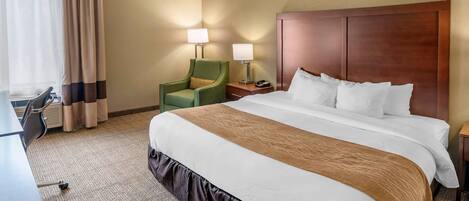 A hotel room with a large bed, two bedside lamps, a green armchair, and a desk with a chair.
