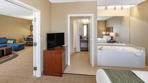 Habitación de hotel con cama, televisor, escritorio, sofá, baño con bañera y un pasillo que conduce a otra habitación.
