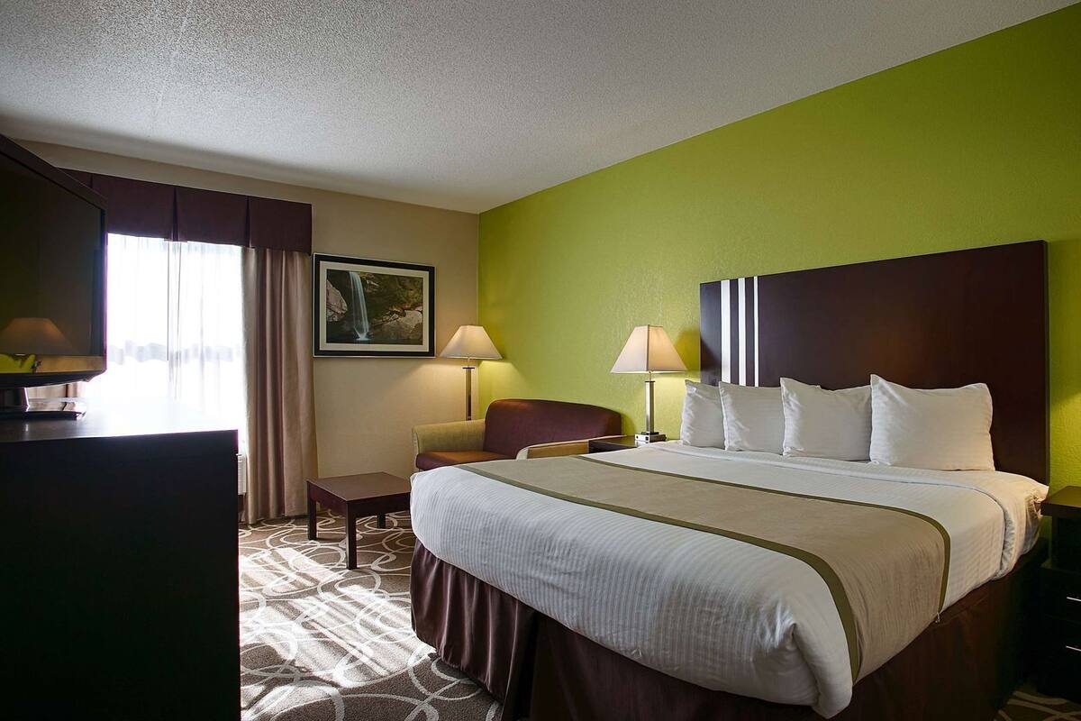 A hotel room with a large bed, two bedside lamps, a desk with a computer, and a window with curtains.