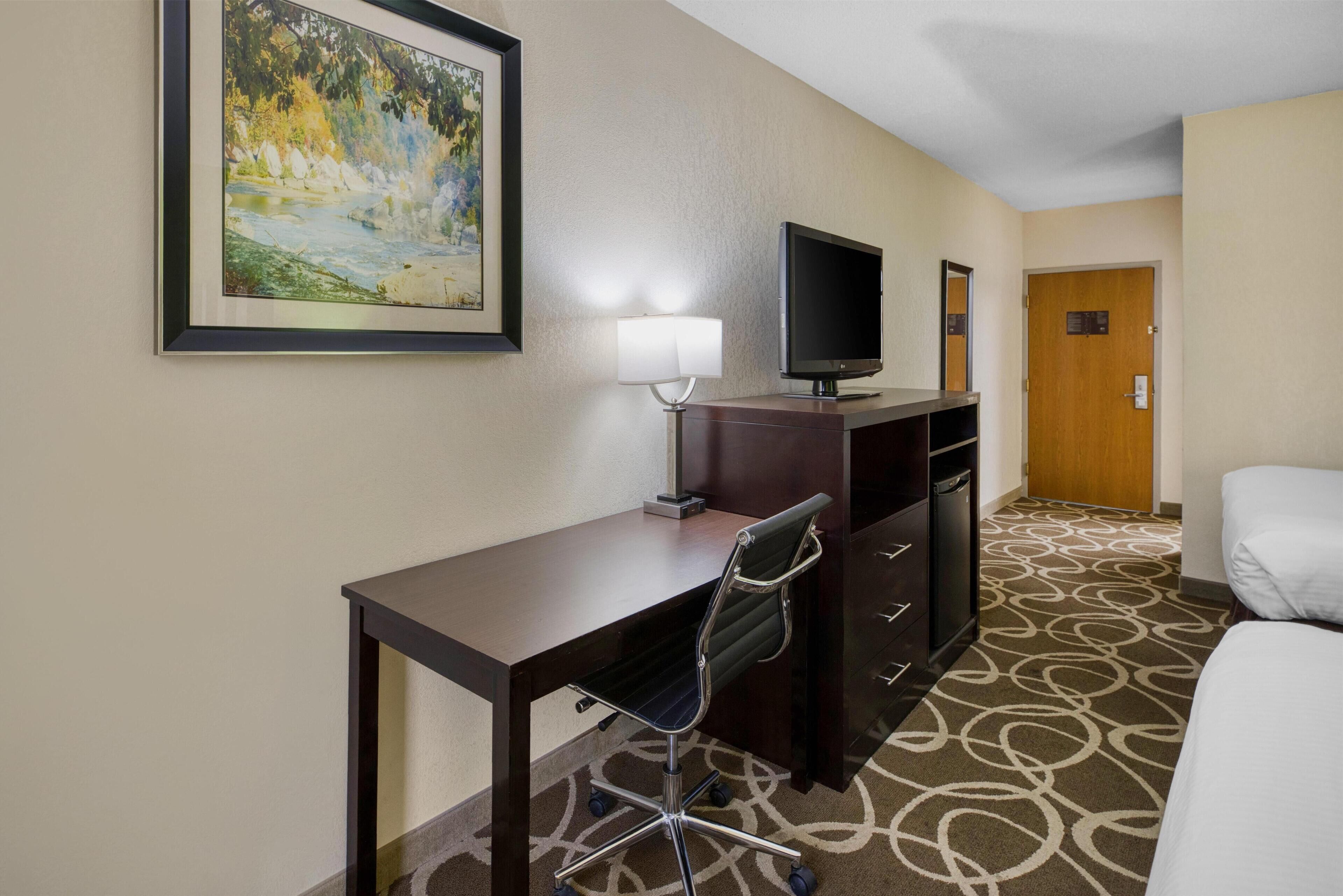 A hotel room with a desk, chair, television, lamp, and a bed.