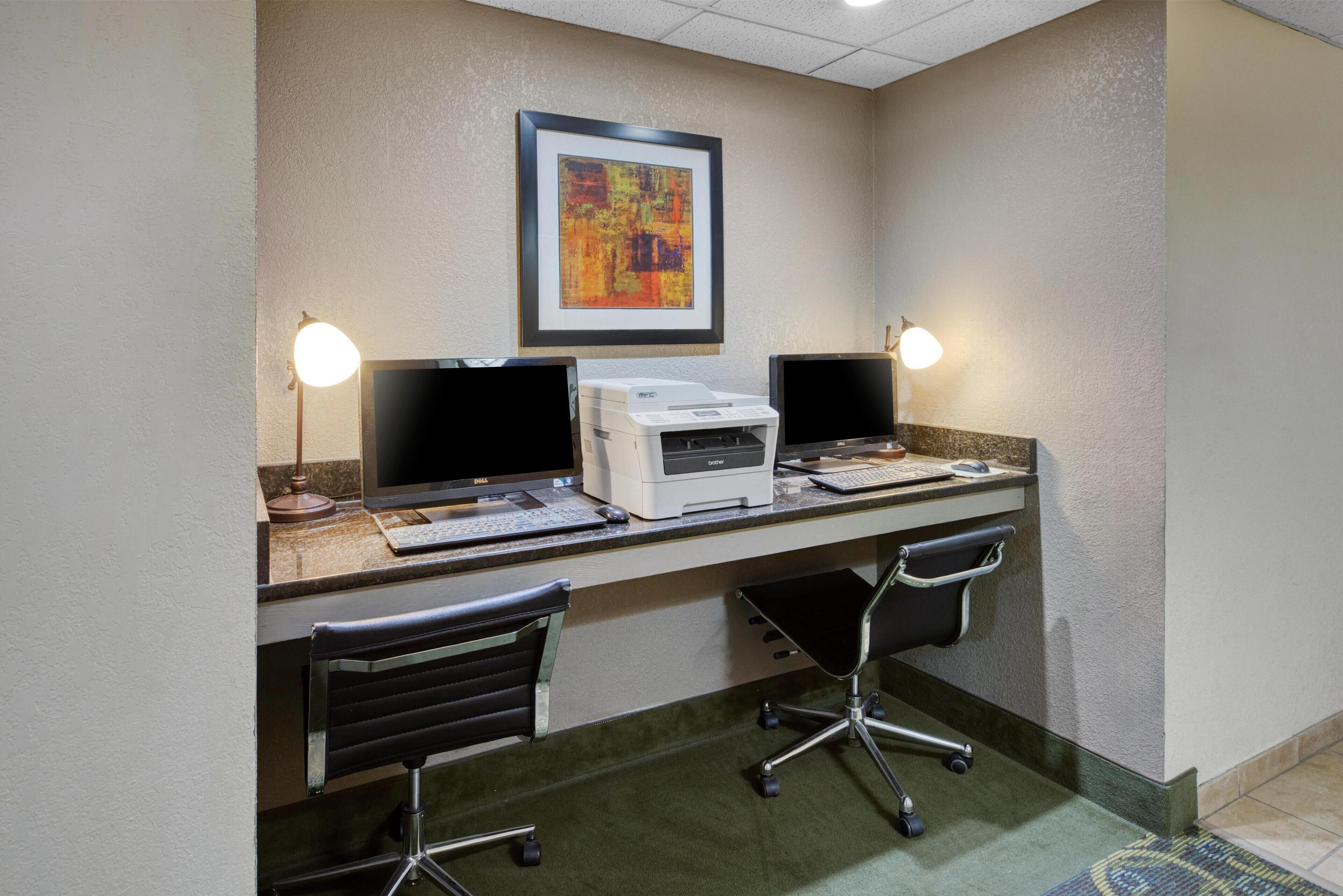 Une chambre d’hôtel équipée d’un bureau, de deux écrans, d’une imprimante et d’un tableau abstrait encadré accroché au mur.