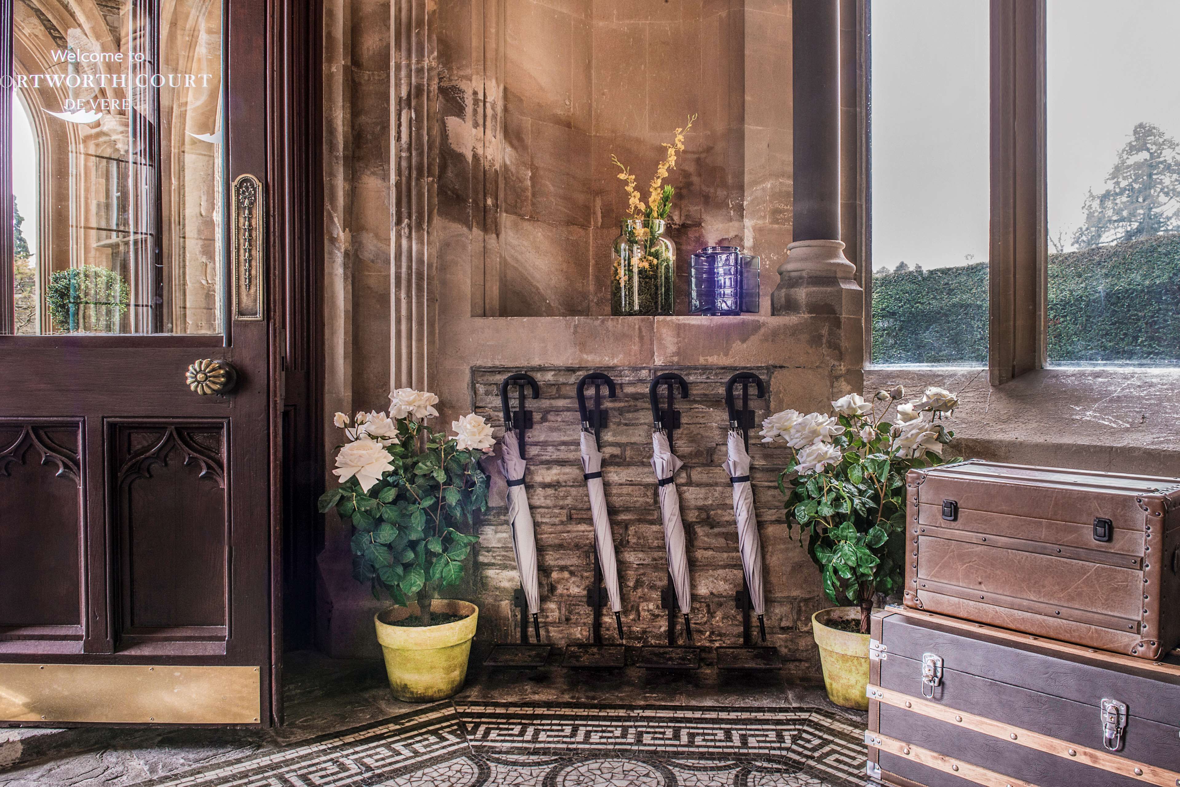 Uma área de recepção com porta de madeira, um banco com guarda-sóis e plantas em vasos.