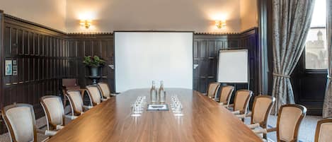 A conference room with a long wooden table, multiple chairs, a projector screen, and two wall-mounted lamps.