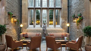 A cozy courtyard with a stone wall, a large window with multiple panes, and a statue in the center. There are four brown leather chairs and a sofa arranged around a small table. - De Vere Tortworth Court (Wotton-under-Edge)