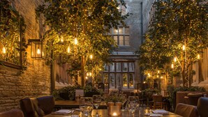 A restaurant with a long table set for dining, surrounded by brick walls and trees adorned with string lights. - De Vere Tortworth Court (Wotton-under-Edge)