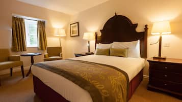 Habitación de hotel con una cama grande, dos mesitas de noche con lámparas, una silla, una mesita pequeña y un ventanal con cortinas.