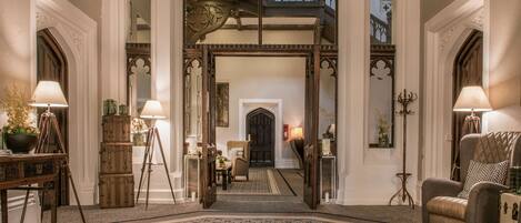 An ornate entryway with a patterned carpet, a grand staircase, and a seating area with a lamp and a chair.