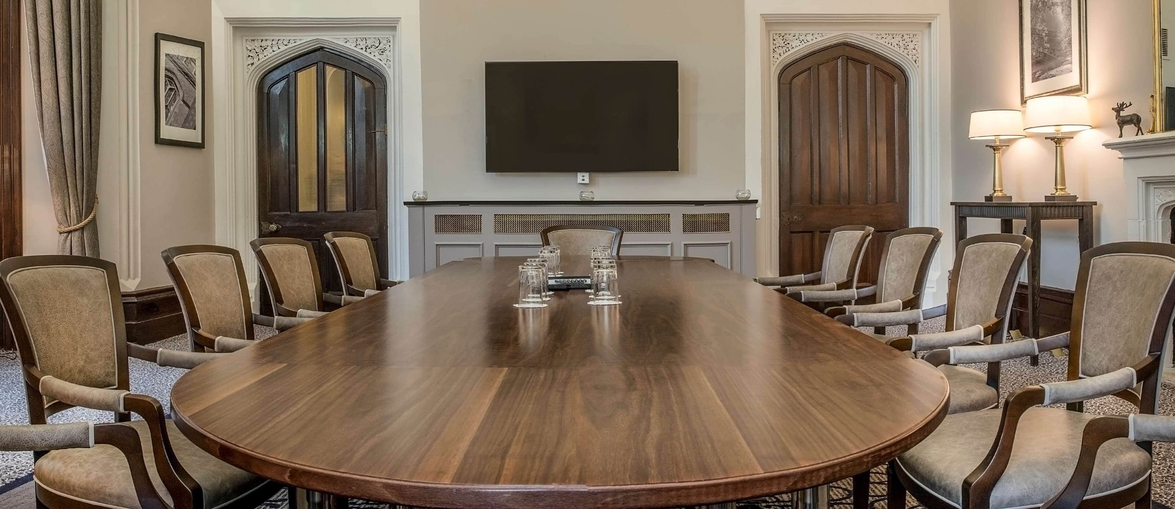 A conference room with a large wooden table, beige upholstered chairs, a flat-screen TV, and a fireplace with a mirror and lamp.