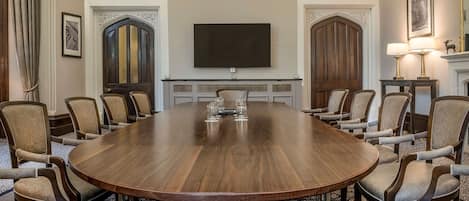 A conference room with a large wooden table, beige upholstered chairs, a flat-screen TV, and a fireplace with a mirror and lamp.