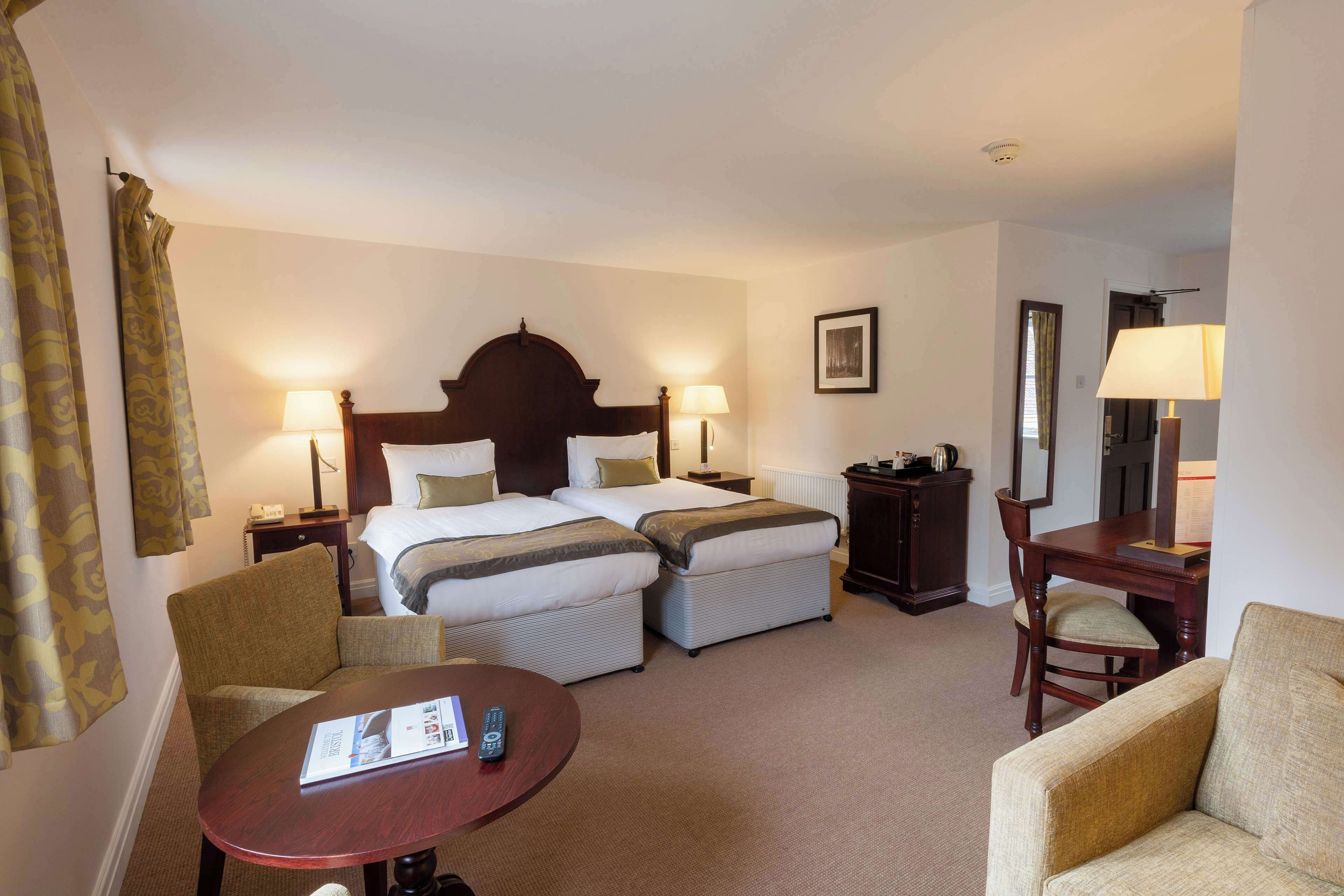 Ein Hotelzimmer mit einem großen Bett, zwei Nachttischen inklusive Lampen, einem Schreibtisch mit Stuhl, einem kleinen Tisch mit Fernbedienung und einem Spiegel an der Wand.