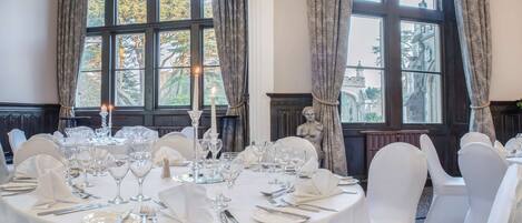 A formal dining room with a table set for a meal, large windows with curtains, and a statue in the background.