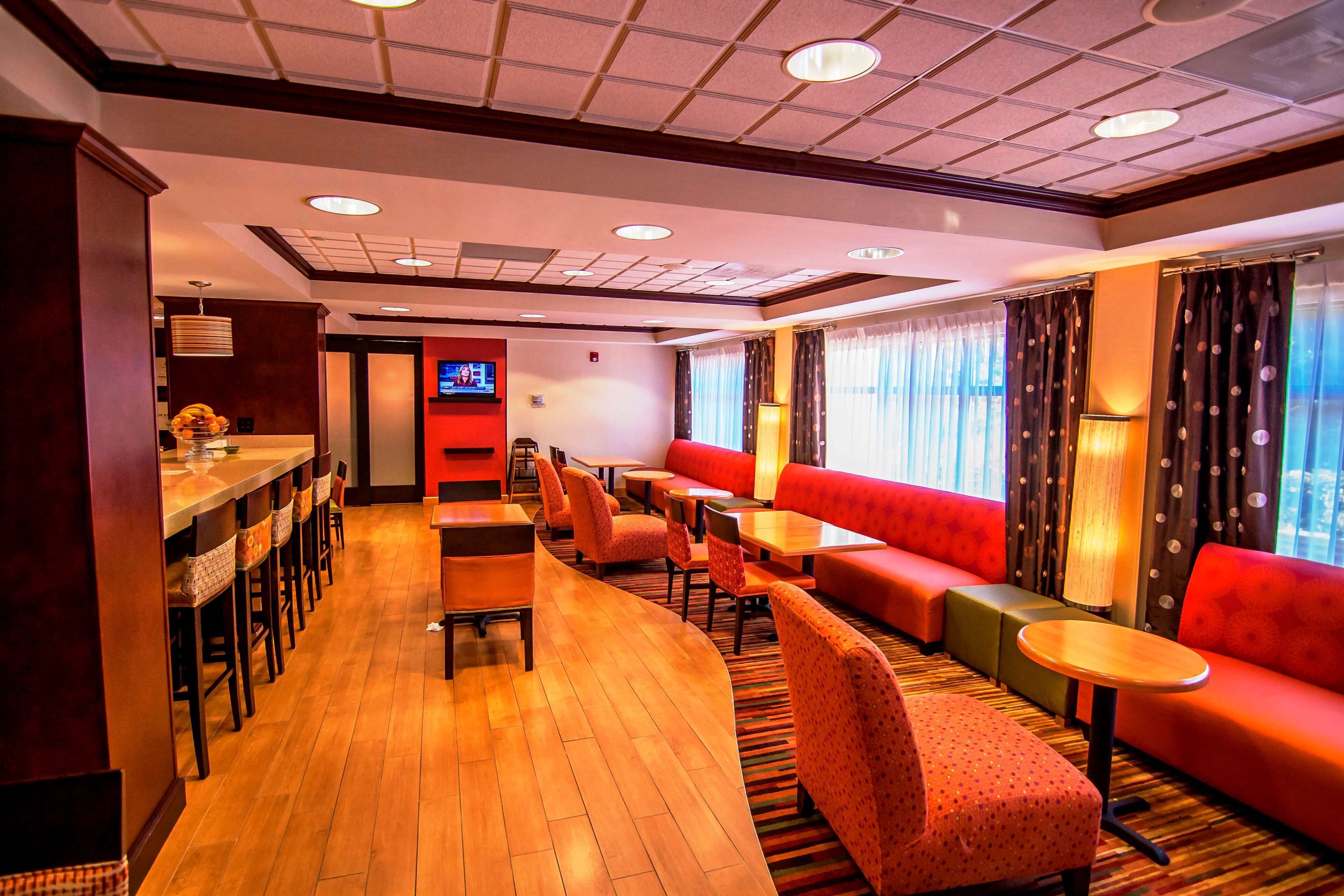 Um hall de hotel com área de bar, disposição de assentos e uma tela de televisão.