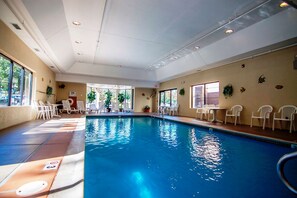 An indoor swimming pool with clear water, surrounded by white chairs and tables, and large windows allowing natural light.