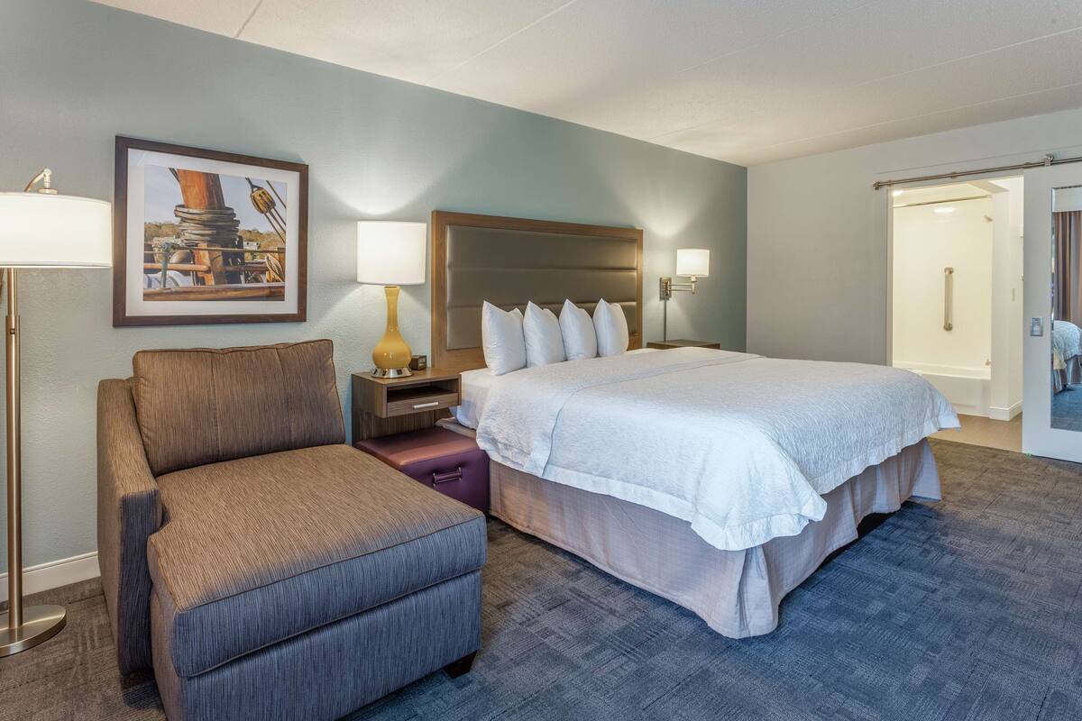 Habitación de hotel con una cama grande, un sofá, una mesita de noche, una lámpara y un cuadro en la pared.
