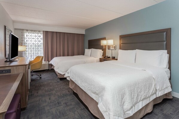 Habitación de hotel con dos camas, un escritorio, una silla, un televisor y un ventanal con cortinas.