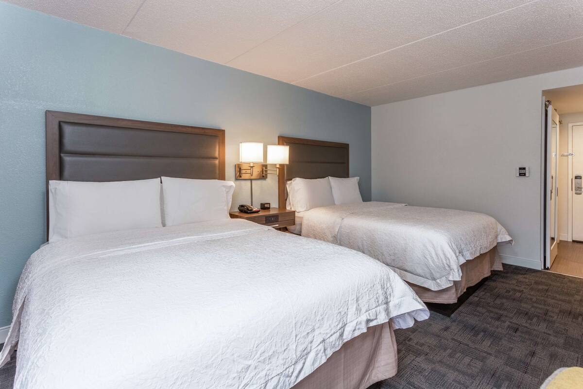 Habitación de hotel con dos camas, una mesita de noche con lámpara y una puerta que da a otra habitación.