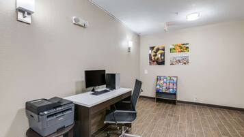 A hotel room with a desk, computer, printer, chair, and a bulletin board with pictures.