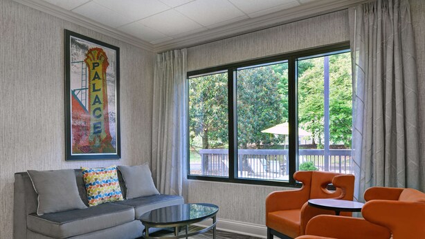 Une chambre d’hôtel avec un canapé gris, une petite table, des chaises orange et une grande fenêtre donnant sur des arbres et une terrasse.