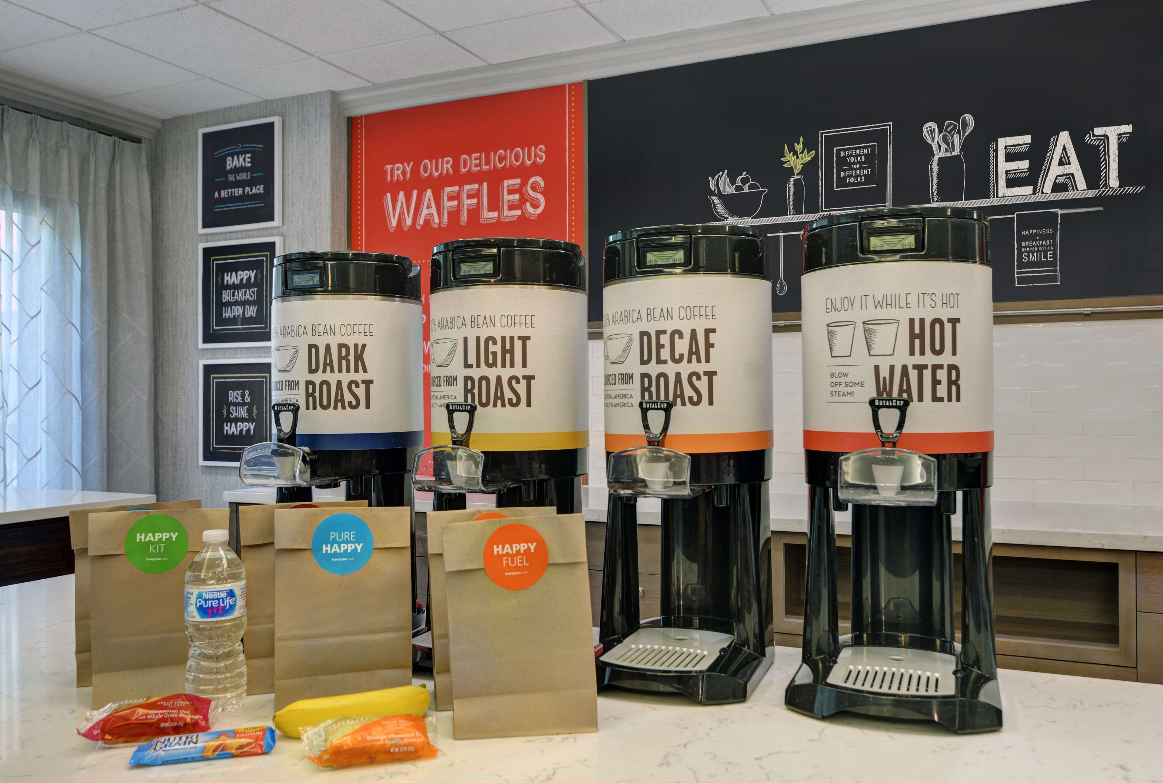 A coffee station with four coffee dispensers labeled 'Dark Roast,' 'Light Roast,' 'Decaf Roast,' and 'Hot Water.' There are also paper bags with 'Happy Fuel' branding and a water bottle.