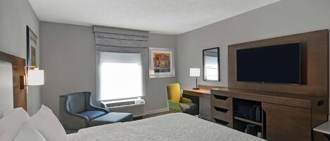 A hotel room with a large bed, a desk with a flat-screen TV, a chair, a window with blinds, and a framed picture on the wall.