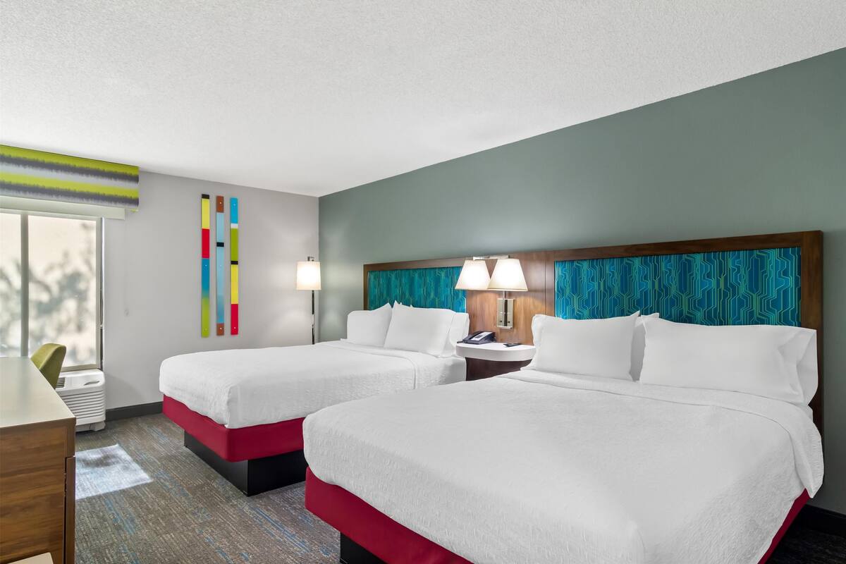 Habitación de hotel con dos camas, un escritorio, una silla, una lámpara y una ventana.
