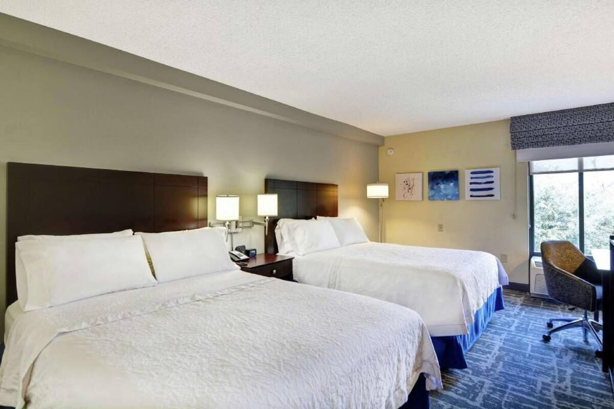 Habitación de hotel con dos camas, un escritorio y una silla. Incluye lámparas de pared y obras de arte.