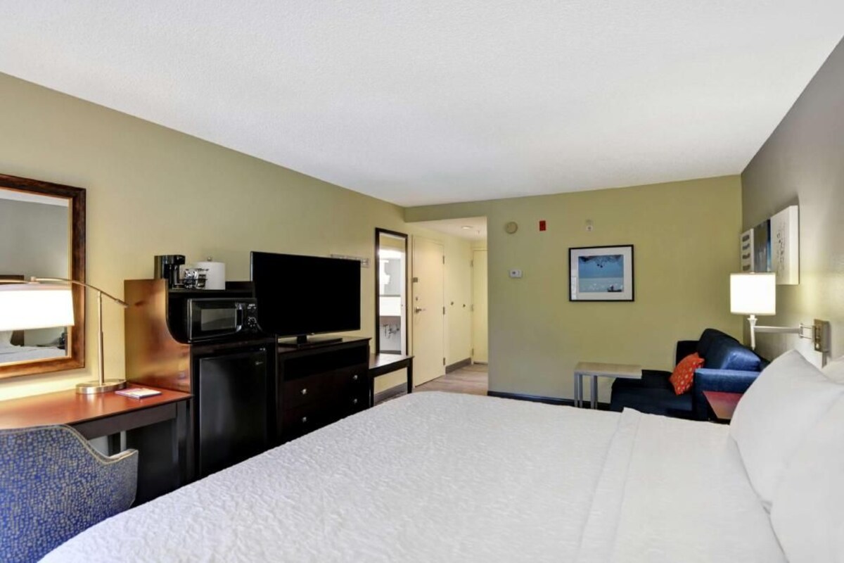 Una habitación de hotel con cama, escritorio con lámpara, televisor, microondas, una mesita, una silla y un sofá.