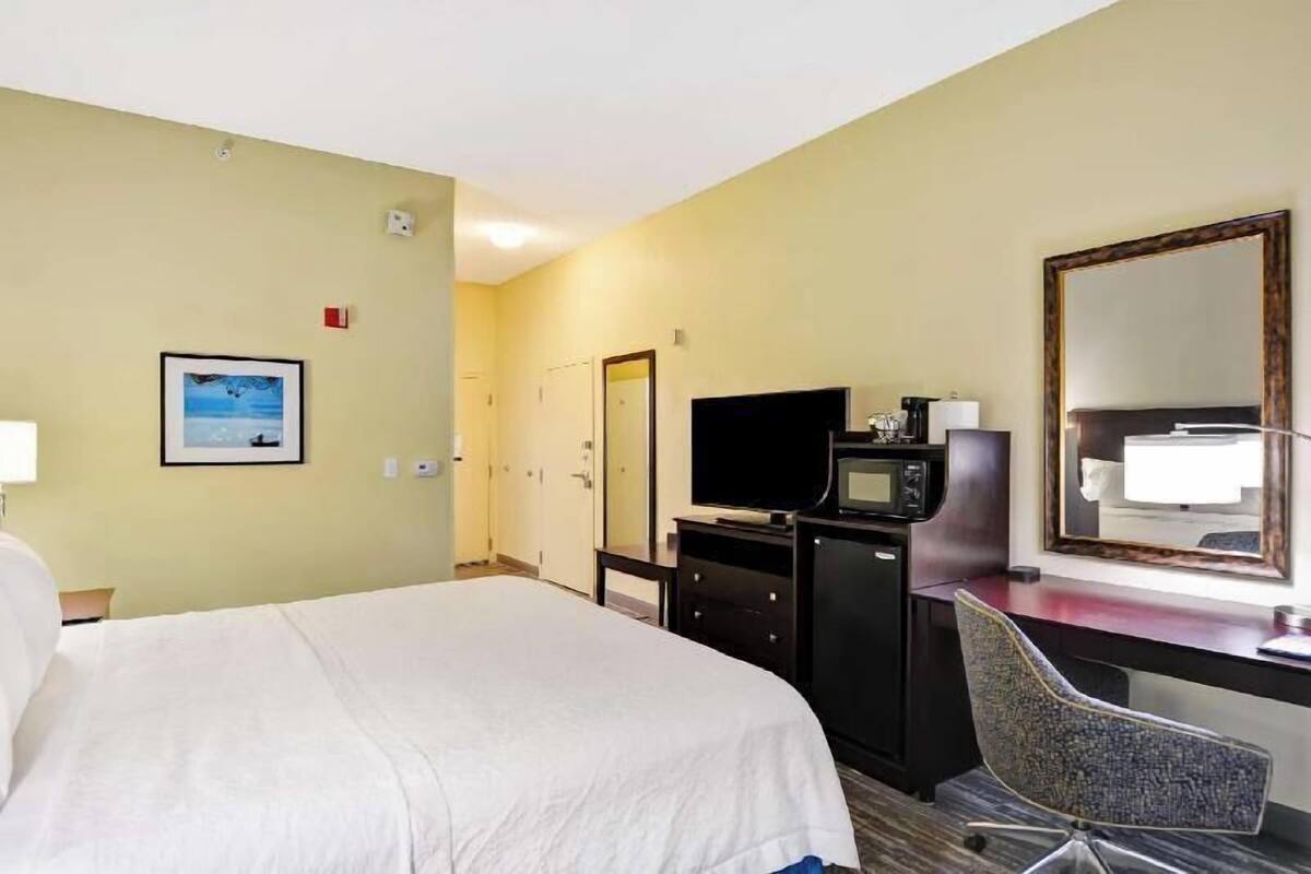 Habitación de hotel con cama, escritorio con televisor, una silla, lámpara de escritorio y un cuadro en la pared.