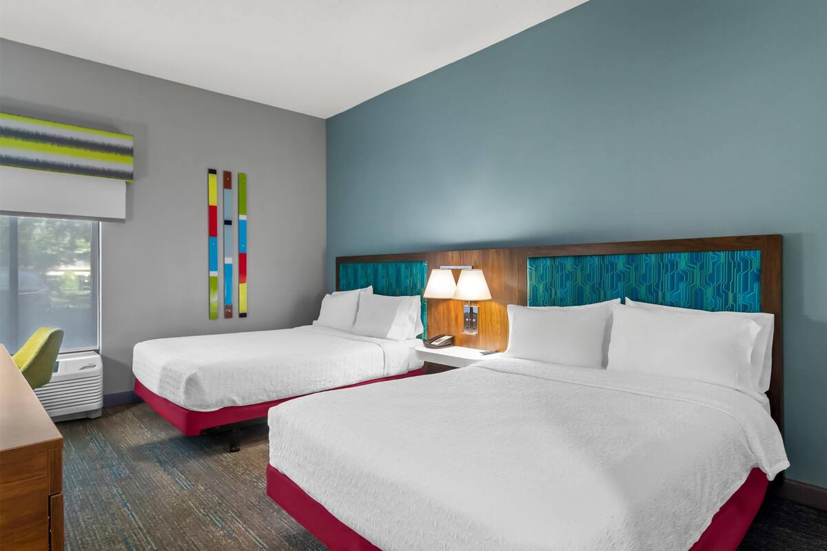 Habitación de hotel con dos camas, un escritorio, una silla, un ventanal y una pared con un diseño abstracto y colorido.