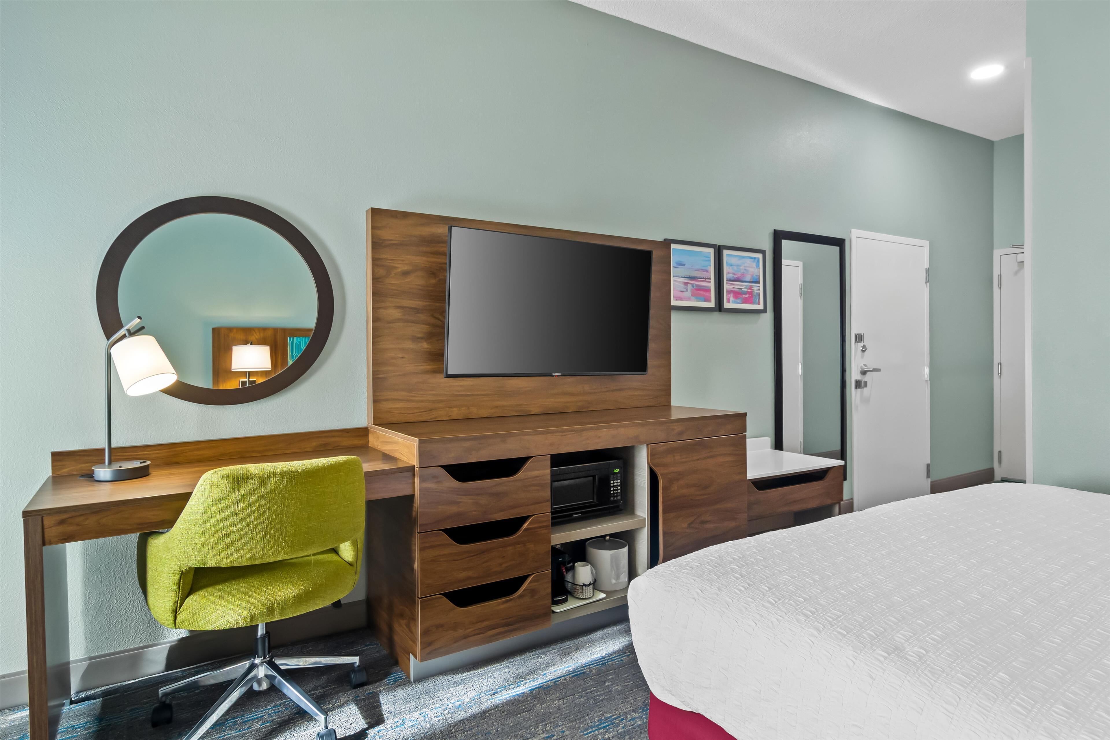 Una camera d'albergo con una scrivania in legno, una sedia verde, una TV a schermo piatto, uno specchio, un letto e un armadio.