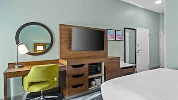 A hotel room with a wooden desk, a green chair, a flat-screen TV, a mirror, a bed, and a wardrobe.