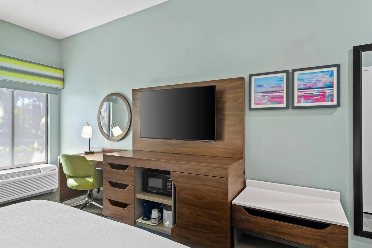 Habitación de hotel con cama, escritorio con televisor de pantalla plana, silla verde, espejo y dos cuadros en la pared.