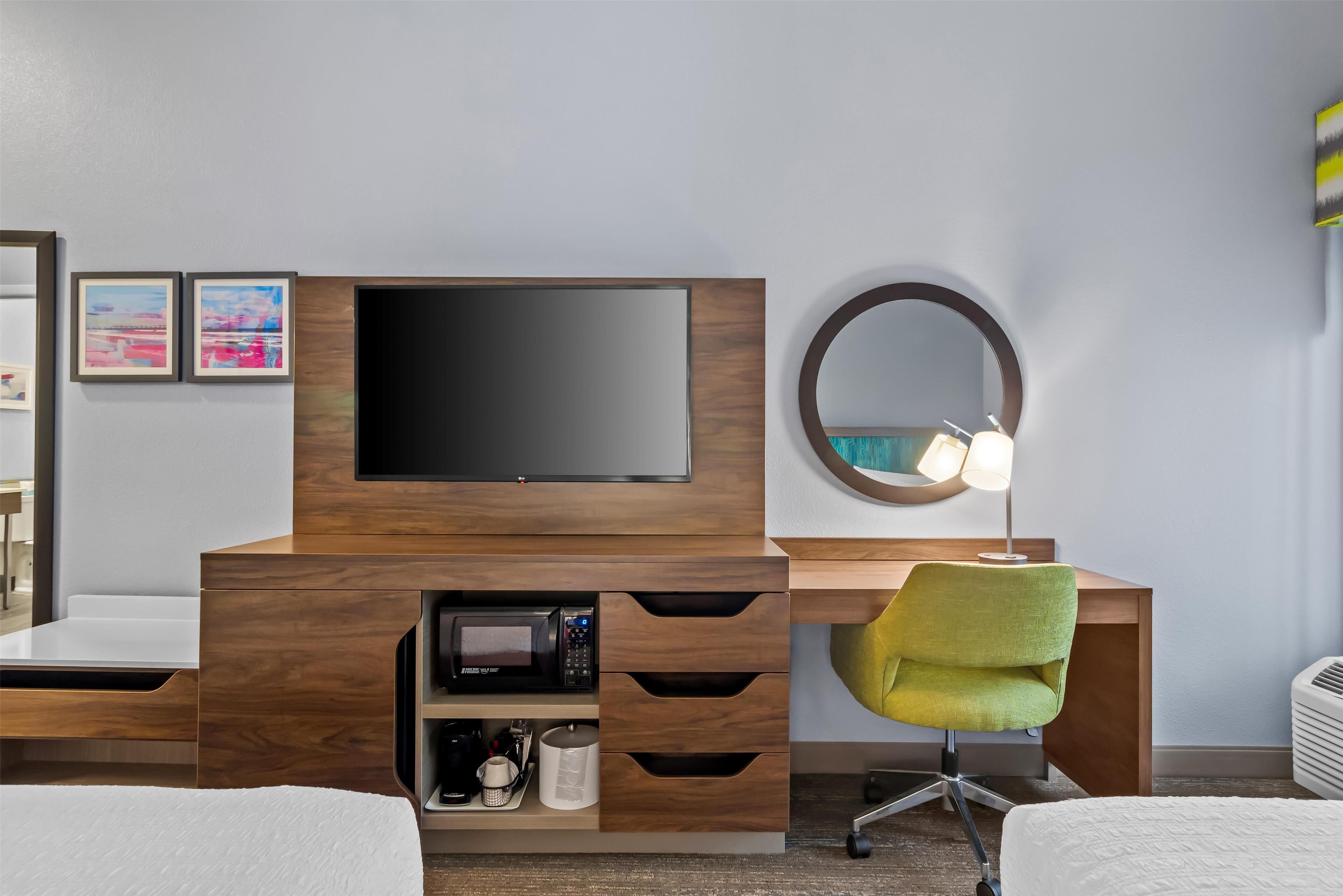 Ein modernes Hotelzimmer mit einem Holzbüro, einem Flachbildfernseher, einem runden Spiegel, einem grünen Sessel und einem Bett.