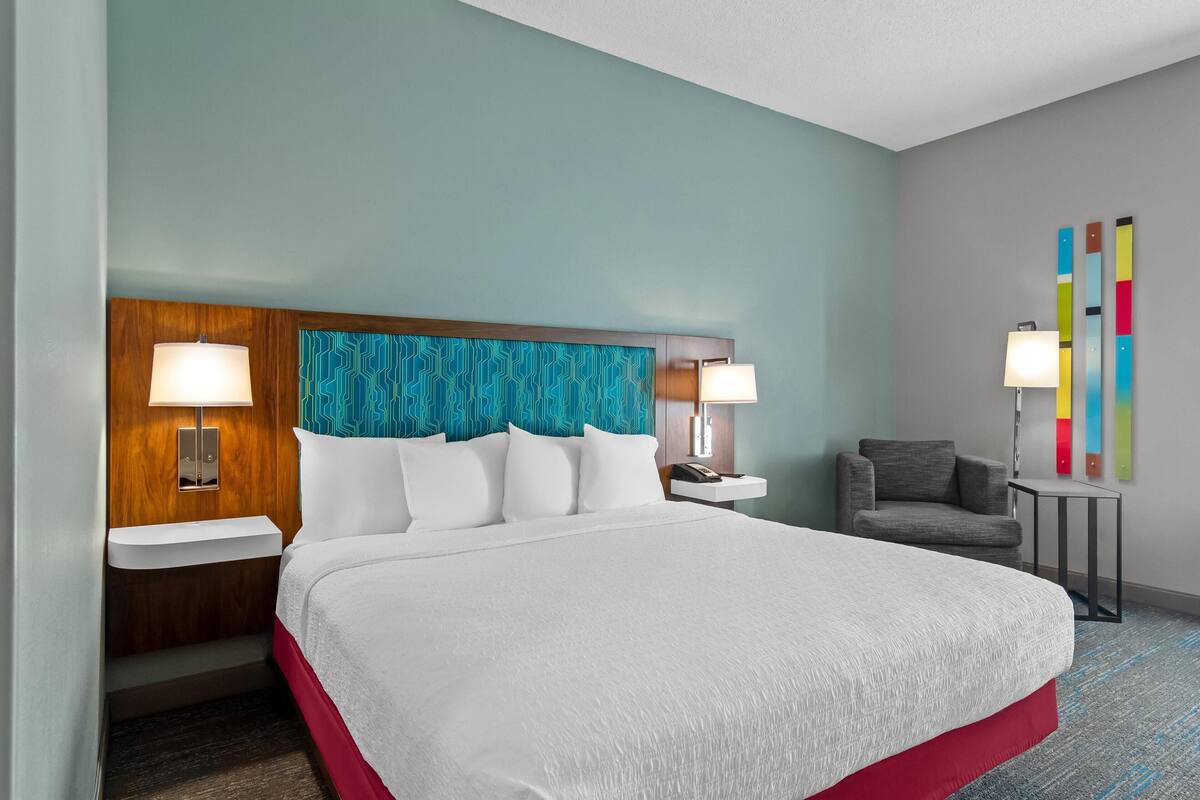Habitación de hotel con una cama grande, dos mesitas de noche con lámparas, un sillón gris y una obra de arte abstracta y colorida en la pared.