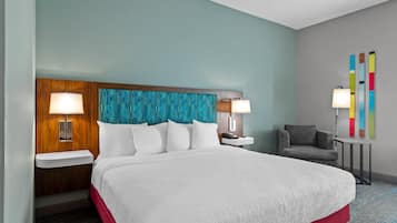 A hotel room with a large bed, two bedside tables with lamps, a gray armchair, and a colorful abstract wall art piece.
