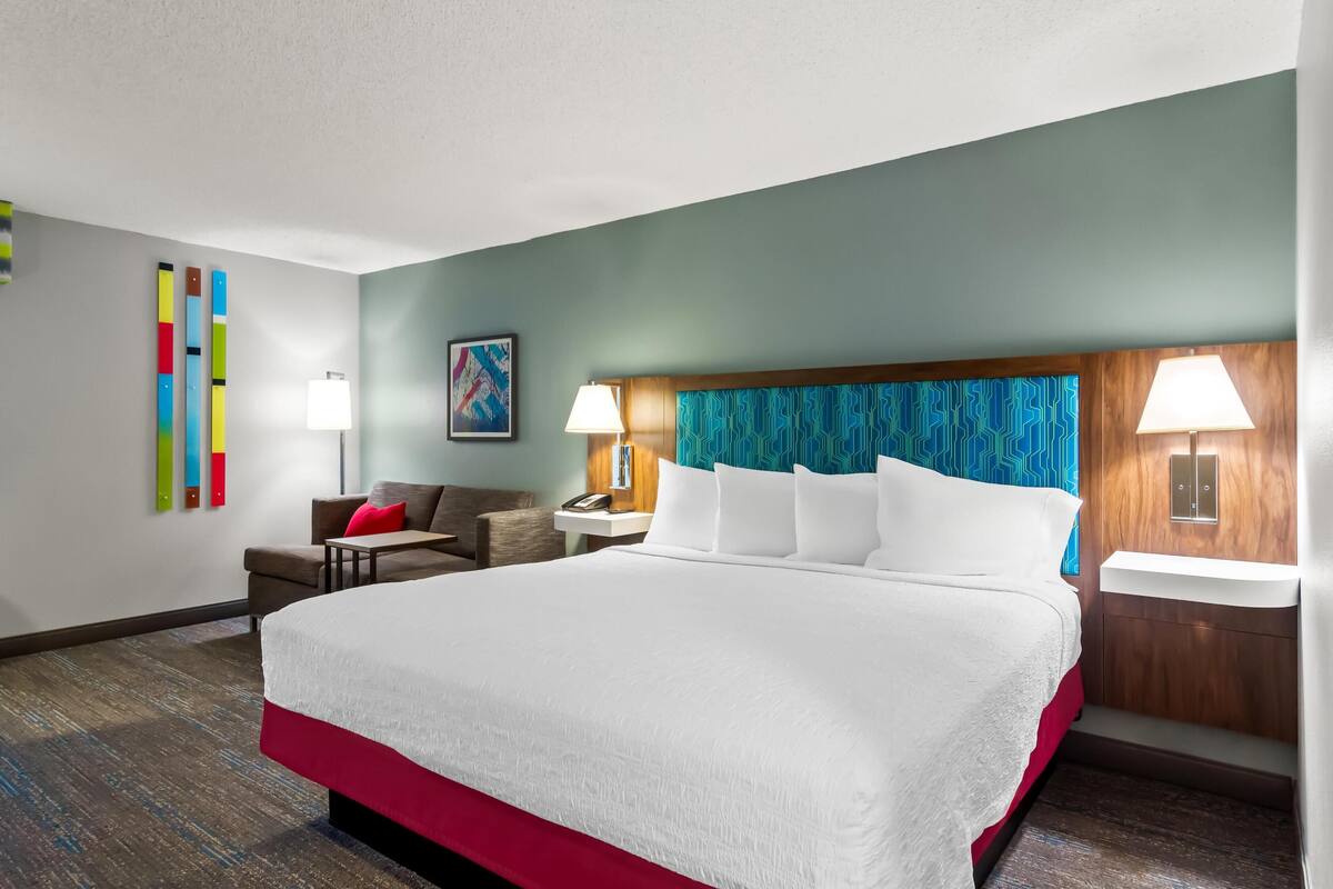 Habitación de hotel con una cama grande, dos mesitas de noche con lámparas, un sofá, una pequeña mesa y un cuadro abstracto de colores en la pared.