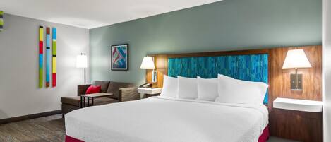 A hotel room with a large bed, two bedside tables with lamps, a sofa, a small table, and a colorful abstract painting on the wall.